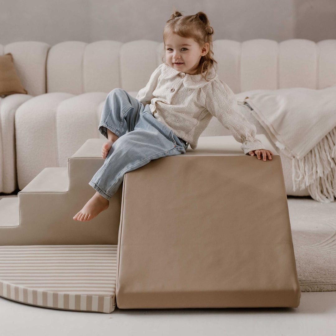 Kind zit bovenaan de beige helling van een HENVAE soft play klimset naast een zachte trap