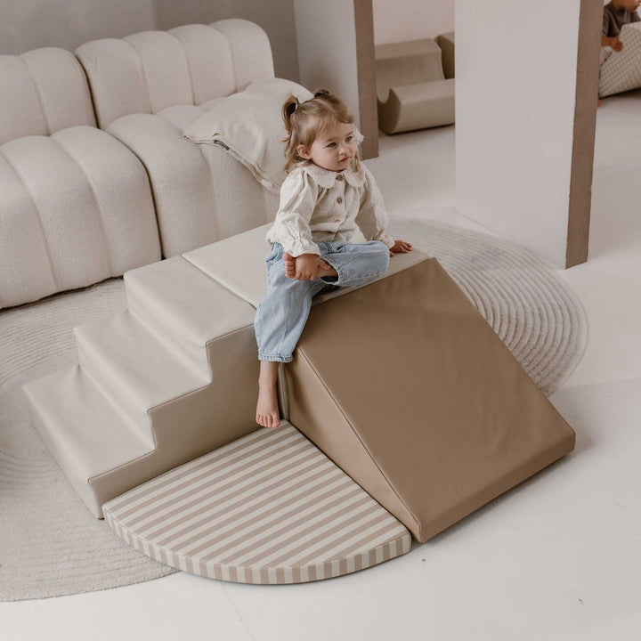 HENVAE beige soft play klimset met trap, glijhelling en gestreepte speelmat, met een kind bovenop